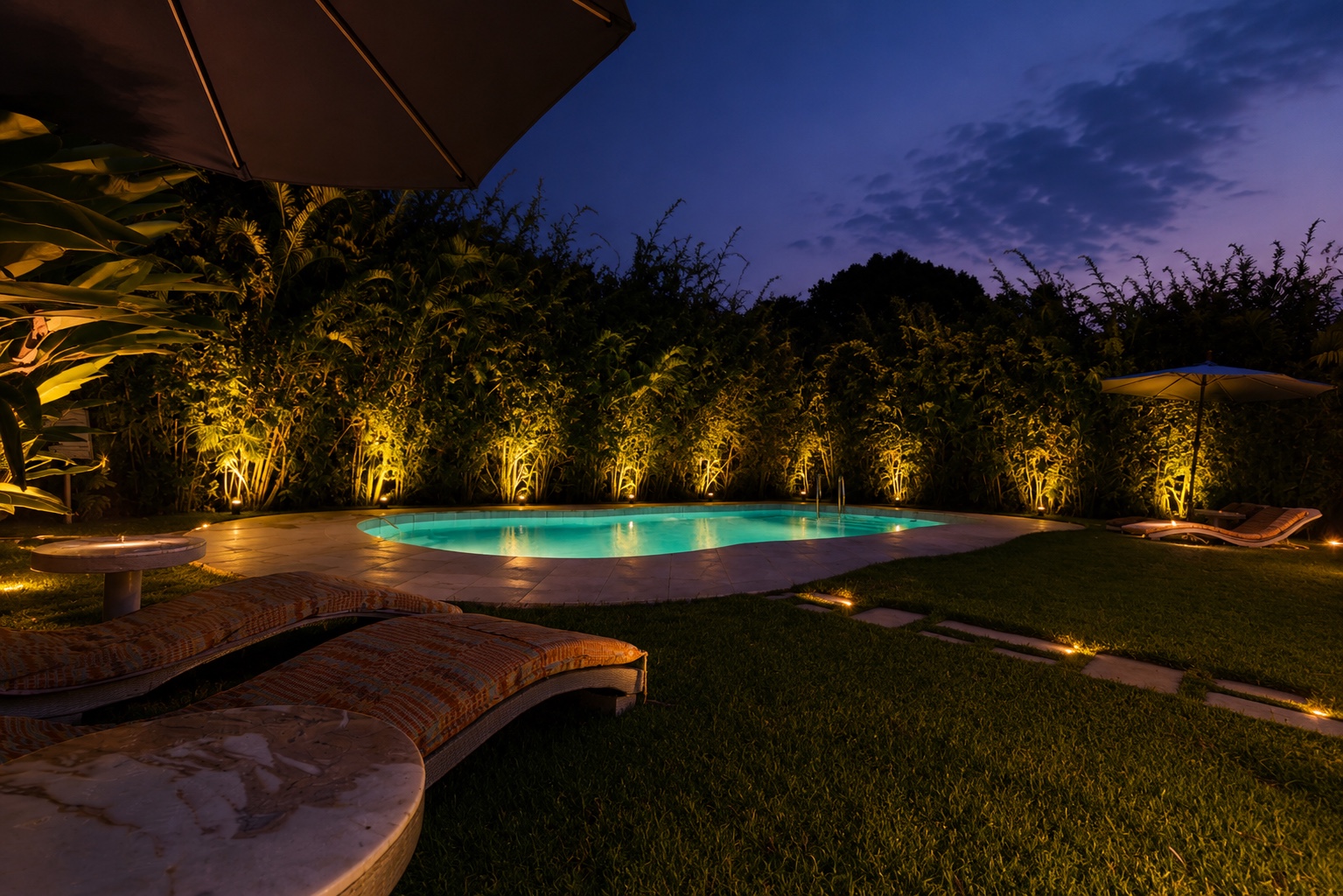 Main pool illuminated at dusk with lit bamboo hedge and loungers