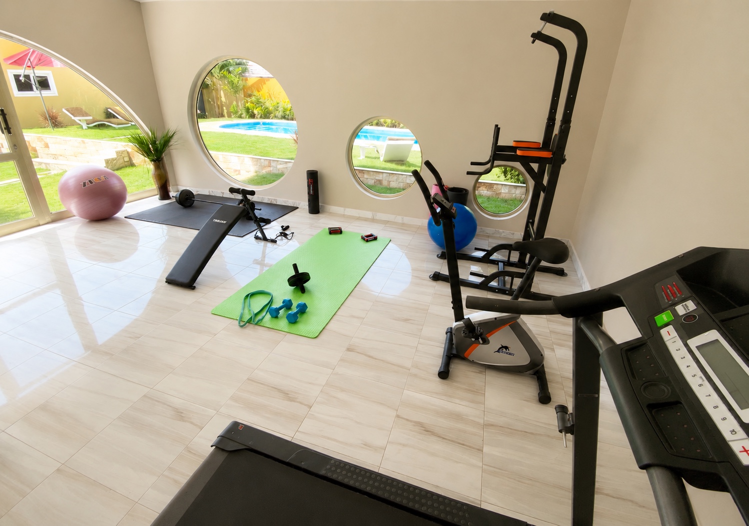 Fitness room with treadmill, weights and round windows overlooking the pool